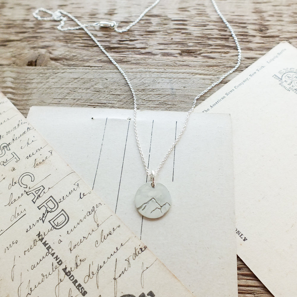 The &quot;Mountains Are Calling Necklace&quot; by Becoming Jewelry, featuring a small silver round pendant engraved with mountain peaks, is displayed on a wooden surface alongside vintage postcards, evoking a sense of adventure and the beauty of nature.