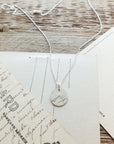 Becoming Jewelry's Mountains Are Calling Necklace on a wooden surface with vintage postcards in the background.