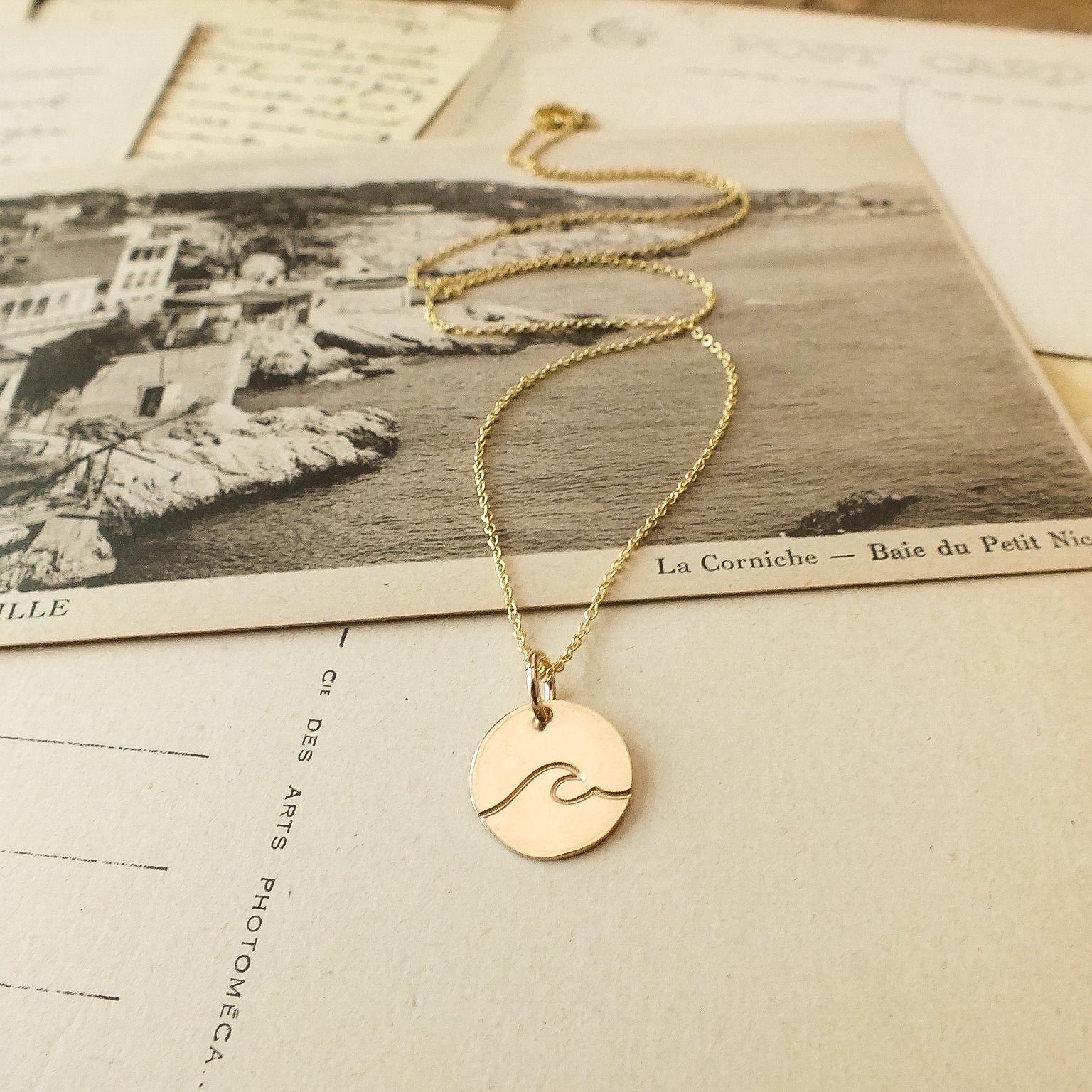 Wave Round Charm Necklace by Becoming Jewelry resting on a vintage postcard featuring a seaside landscape.