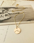 Wave Round Charm Necklace by Becoming Jewelry resting on a vintage postcard featuring a seaside landscape.