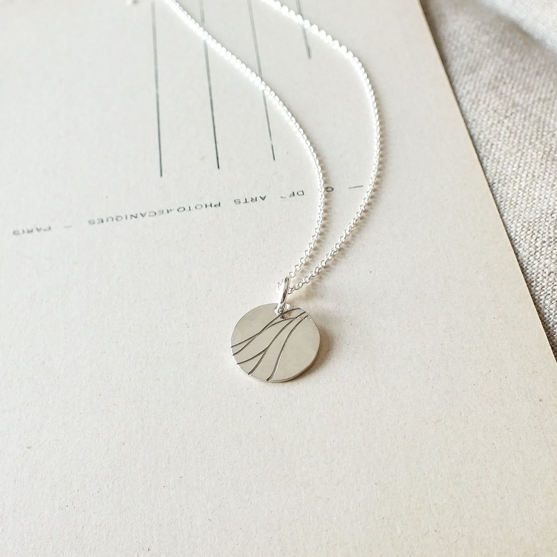 A silver River Necklace by Becoming Jewelry, featuring a round pendant with minimalist line designs, rests on an off-white textured surface with faint lines, symbolizing the indomitable spirit of remarkable women.