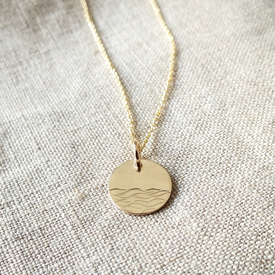 The Sea Necklace by Becoming Jewelry features a gold round pendant engraved with a minimalistic wave pattern, reminiscent of the ocean, displayed on a textured fabric background.