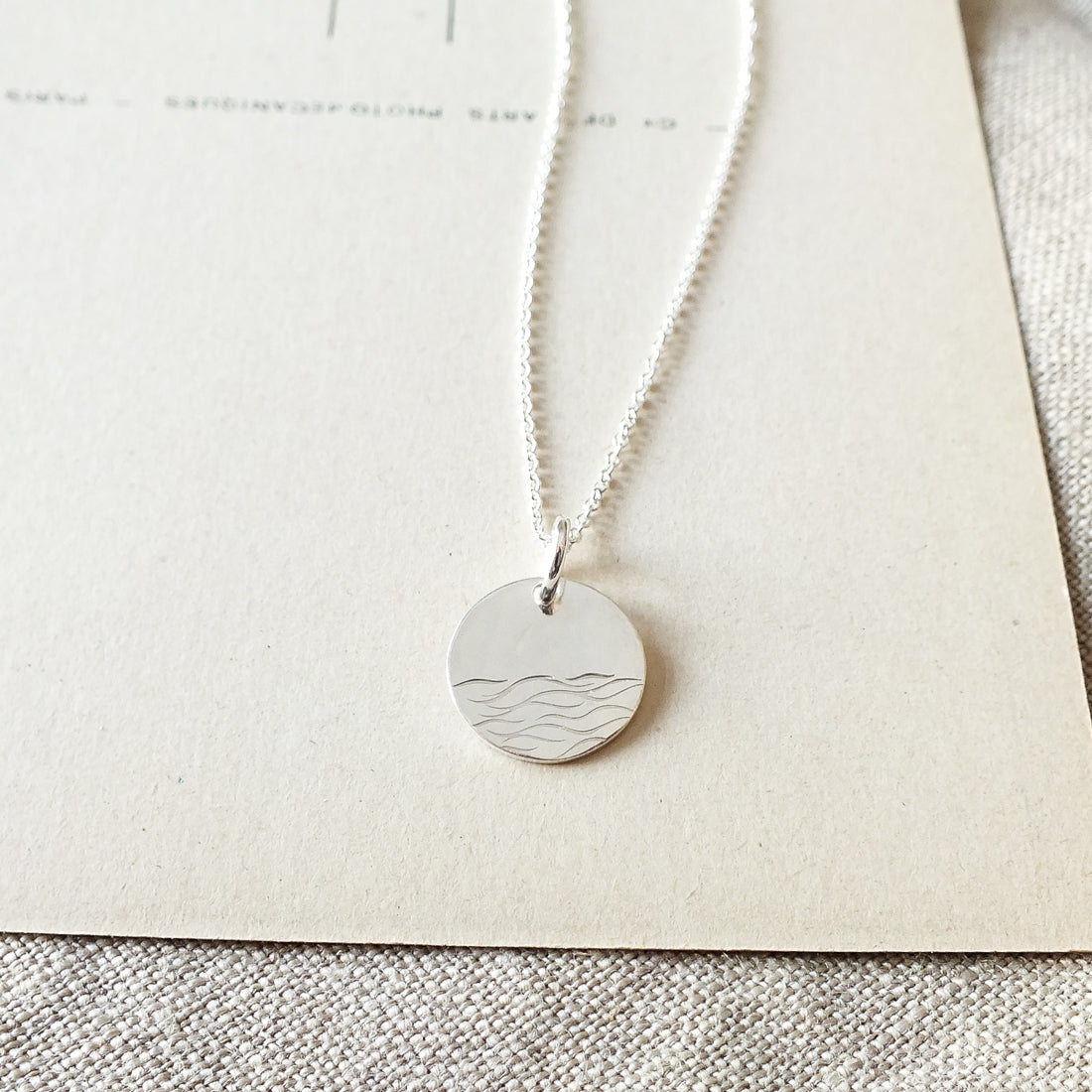 Close-up of The Sea Necklace by Becoming Jewelry, featuring a round pendant with an ocean wave pattern, placed on a beige textured background.