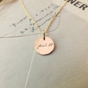 A gold-filled Fuck It Necklace by Becoming Jewelry with a circular pendant inscribed with the phrase "just do it" resting on a paper surface with handwritten text.
