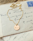 A Loved Necklace by Becoming Jewelry with a circular pendant inscribed with the name "linda" resting on top of an old, handwritten letter.