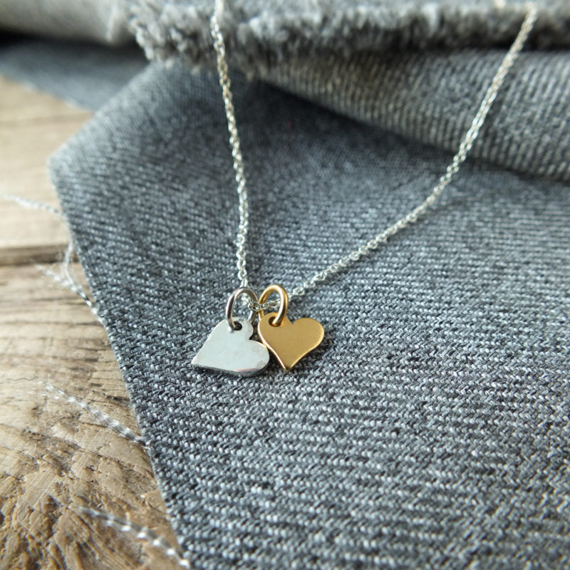A Becoming Jewelry Tiny Hearts Necklace with two heart-shaped pendants resting on a grey fabric surface.